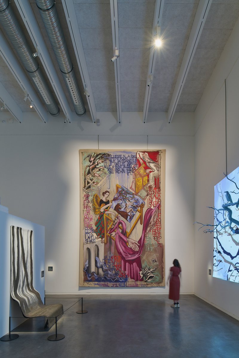 Exhibition room with concrete ceiling panels, exposed ductwork, and a visitor viewing a large tapestry