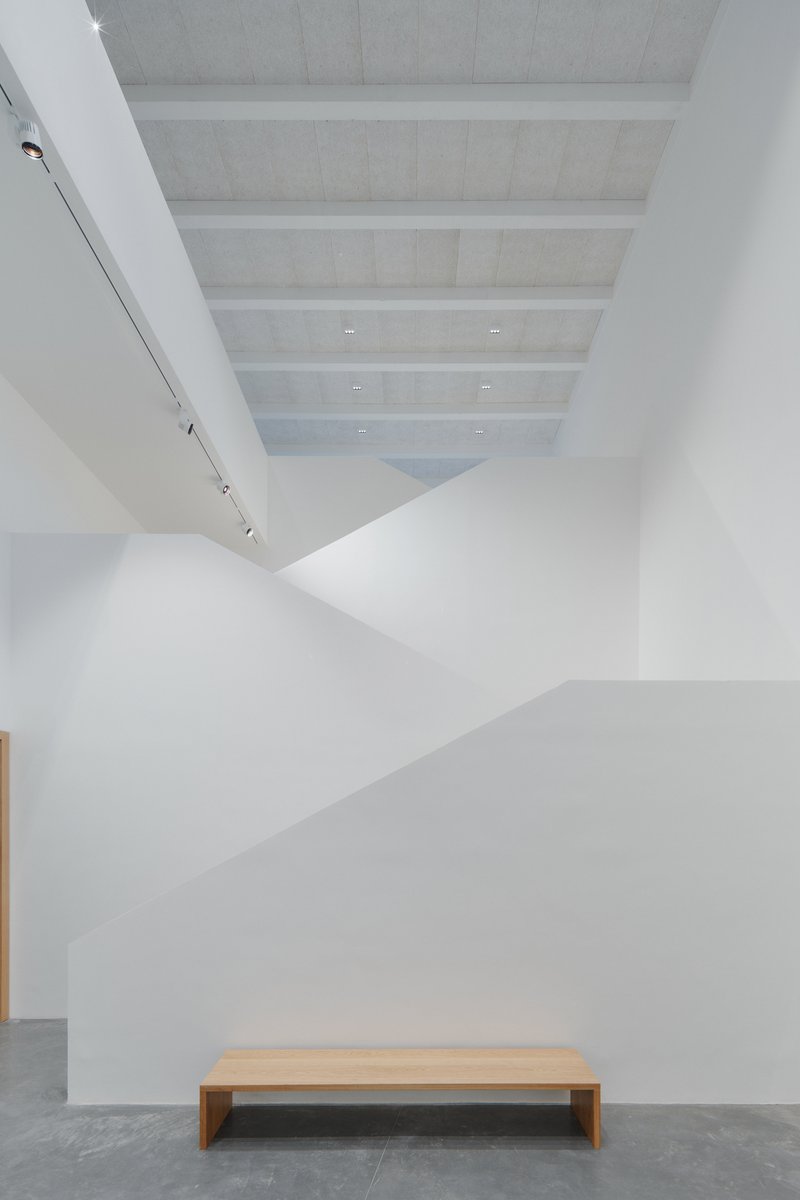White gallery wall with angled ceiling planes meeting above a timber bench on concrete floor