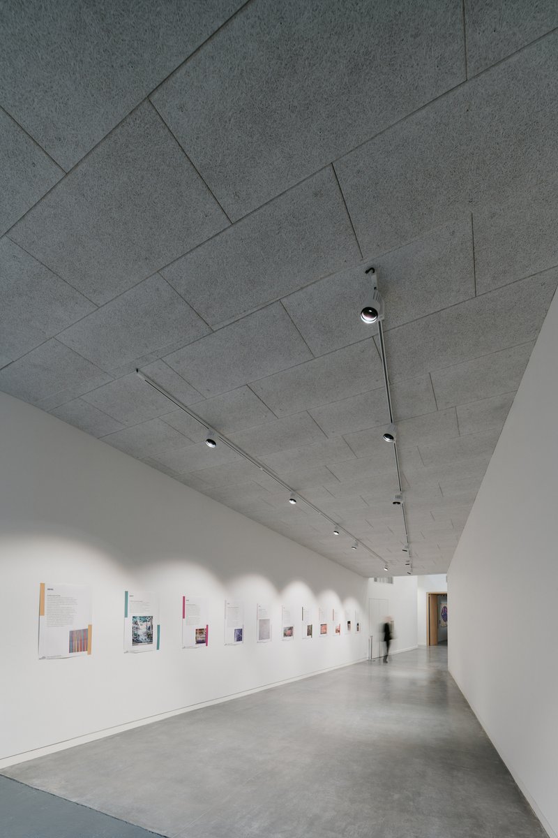 Gallery hallway with rectangular concrete ceiling panels and a figure passing small framed works on white walls
