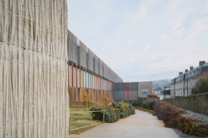 Boardmarked concrete facade alongside a pedestrian path with planted beds on an overcast day