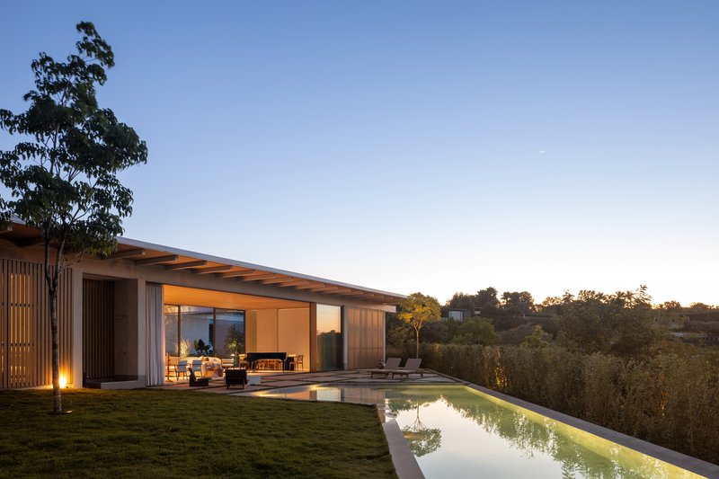 Timber-clad pavilion with overhanging roof and glazed walls beside a lap pool at dusk