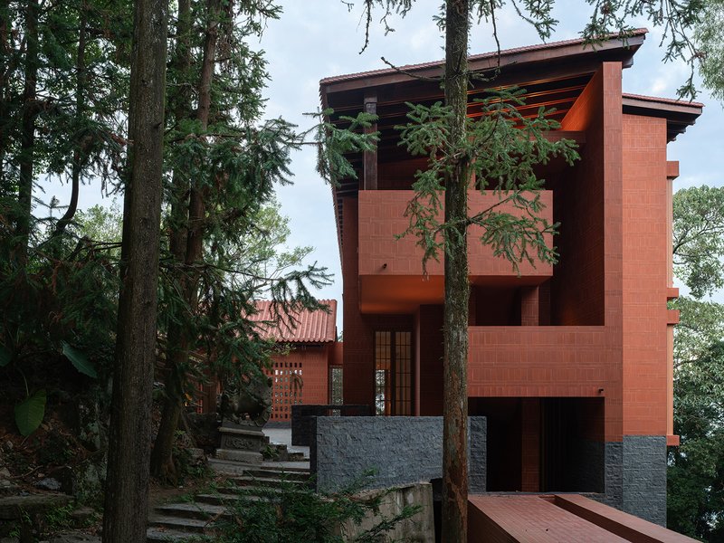 Multi-level red brick volume with exposed timber roof framing rising through tall pine trees