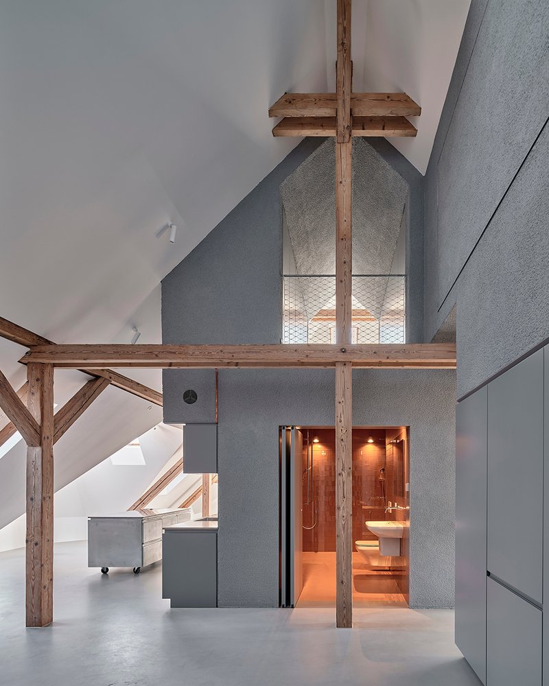 Double-height space with exposed timber beams and view into illuminated bathroom through glass partition