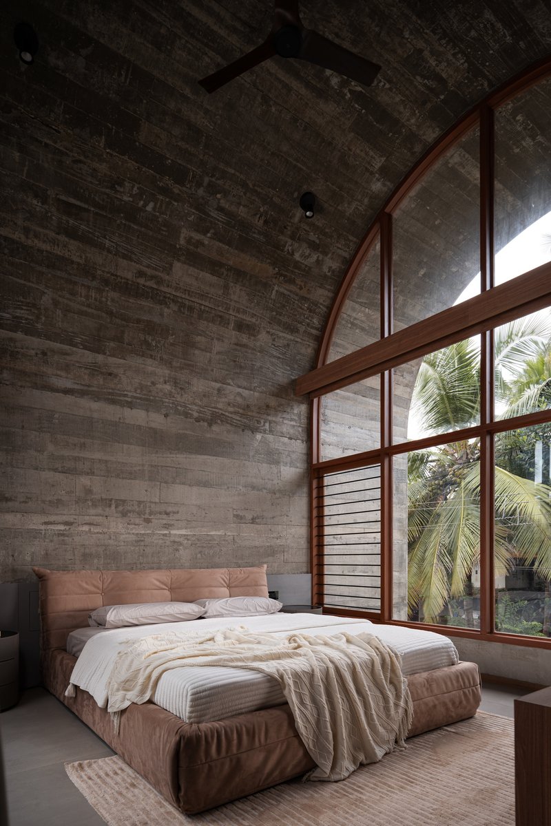Bedroom with arched timber-framed windows overlooking palm trees beneath a board-formed concrete vaulted ceiling