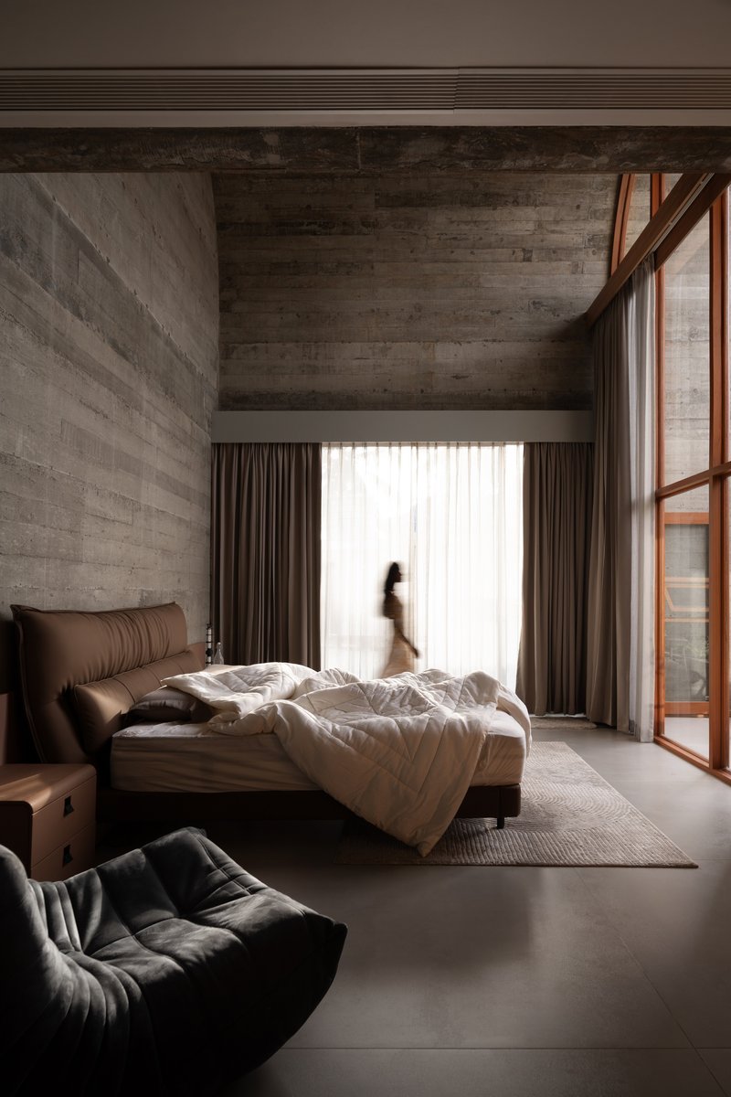 Bedroom with boardmarked concrete walls, tall curtained window, and a figure walking in soft daylight