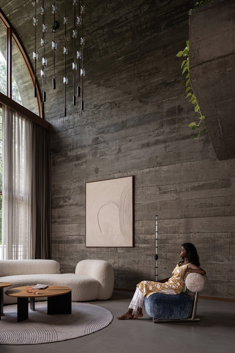 Double-height living space with board-formed concrete wall, pendant light installation and person seated in armchair