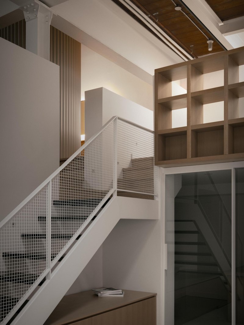 White staircase with mesh railing ascending past timber shelving and glazed doorway in double-height space