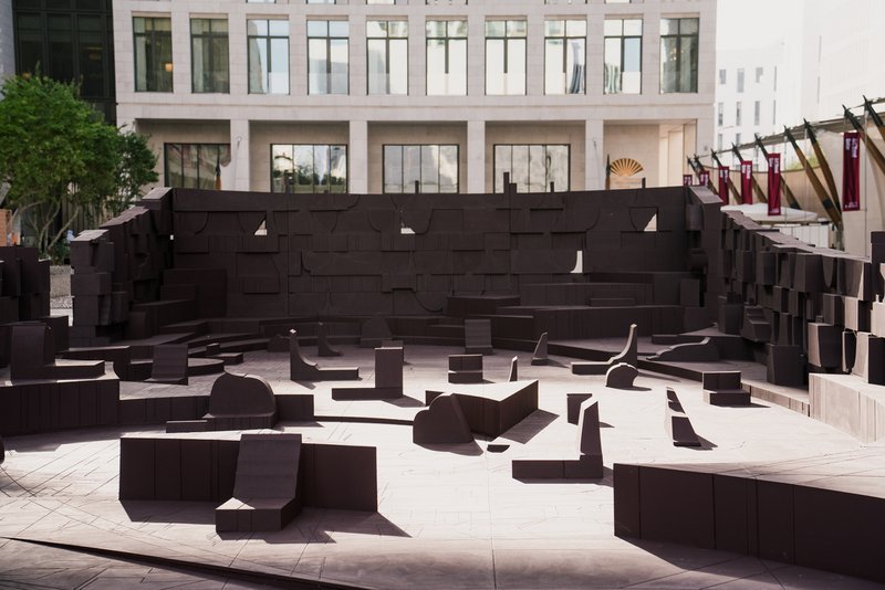 Public plaza with dark brown sculptural seating elements and stepped amphitheater wall beneath surrounding buildings