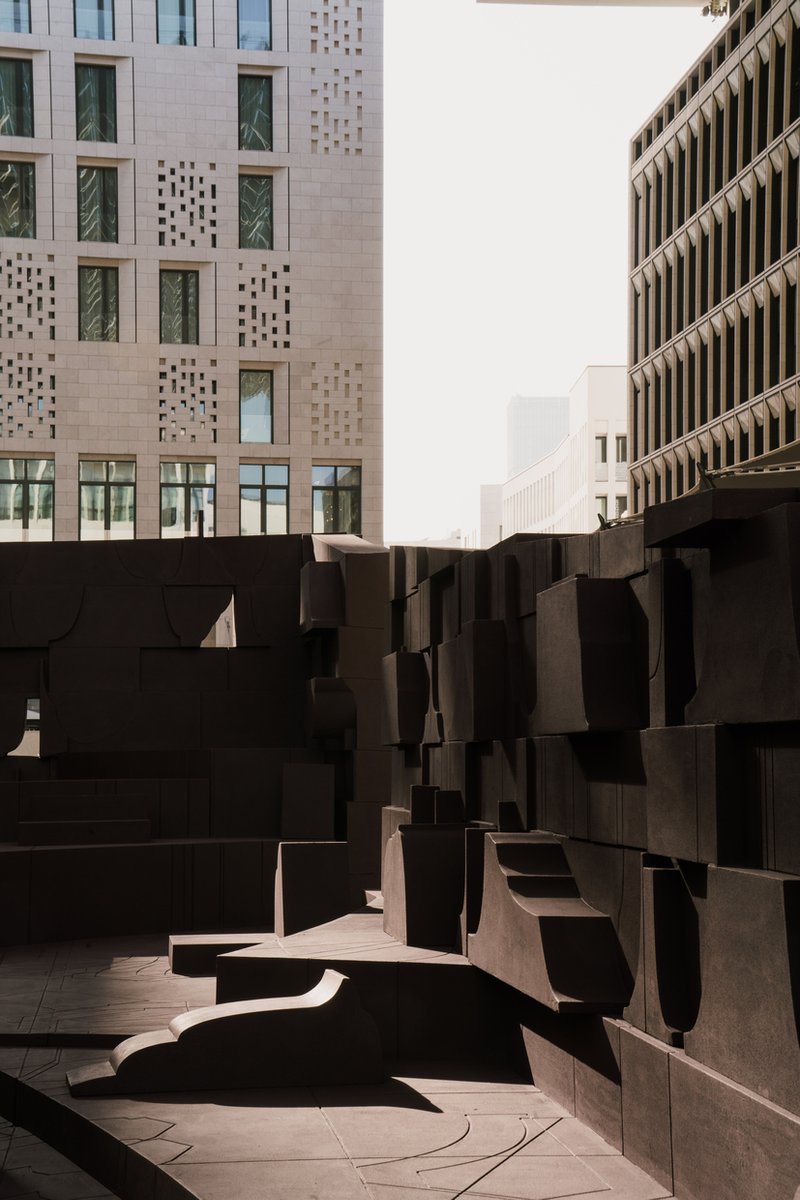 Narrow passage between dark sculpted seating blocks with perforated facade visible beyond in hazy daylight