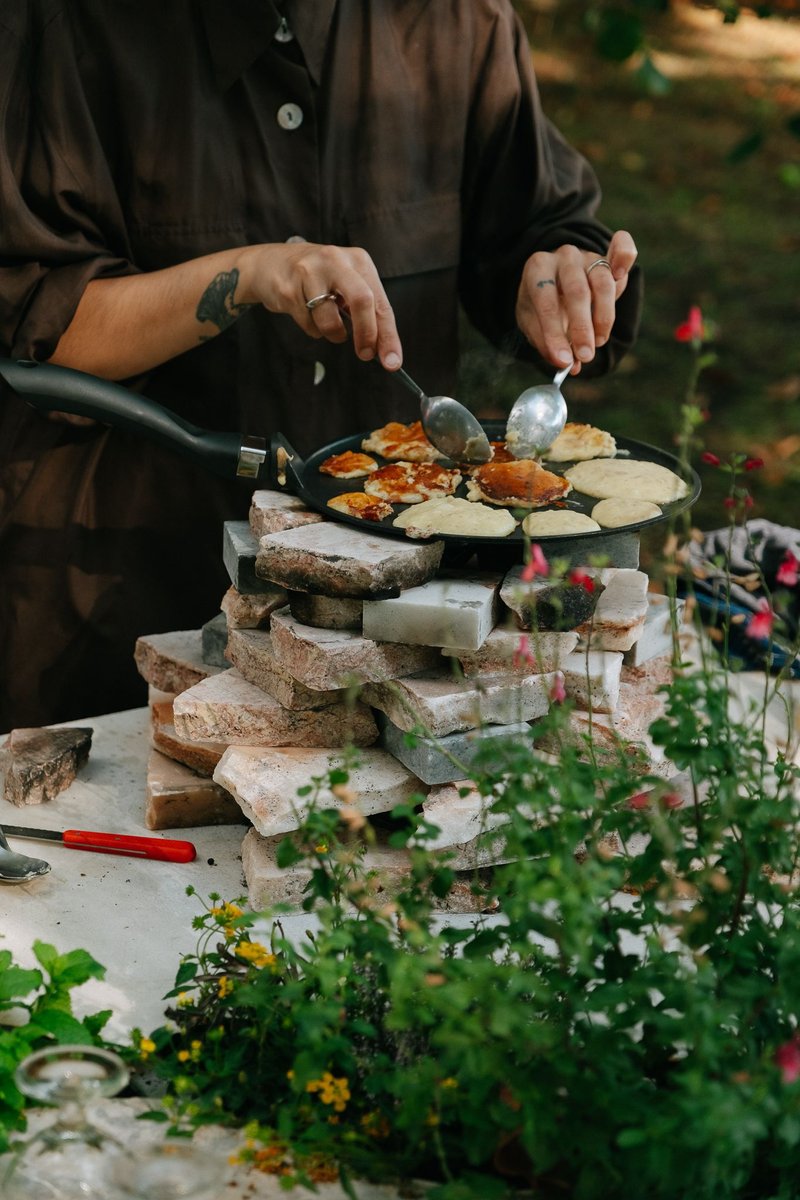 Hands working a flat griddle atop a stacked stone cooking platform surrounded by flowers