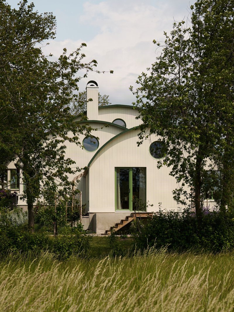 White vertical wood siding and curved green roof with round windows framed by tall trees
