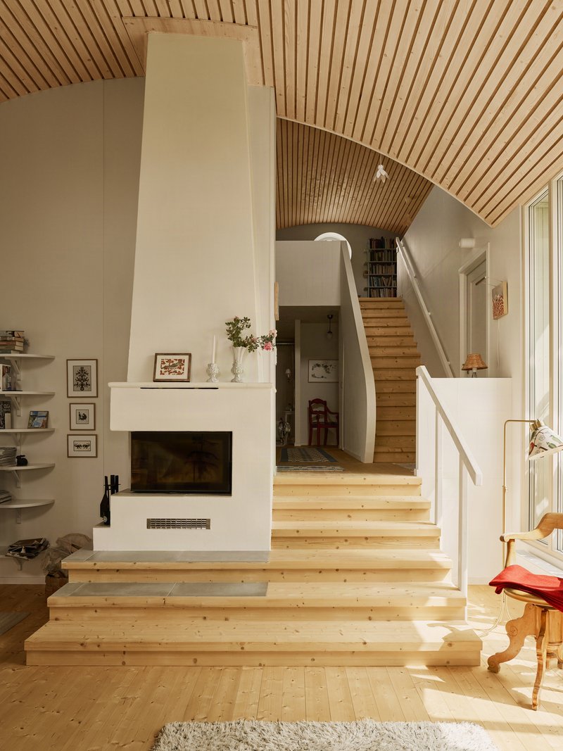 Split-level interior with pale timber stairs rising past a fireplace under curved wood slatted vaulting