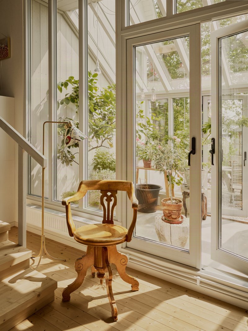 Glazed corner with full-height white-framed doors opening to a greenhouse with potted plants in sunlight