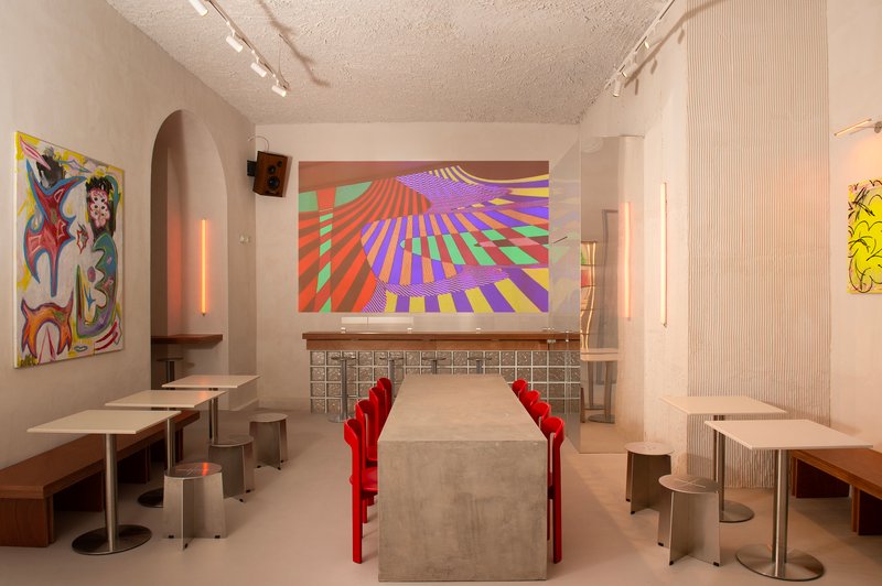 Interior dining area with concrete tables, red chairs, white mosaic tile bar and projected colorful geometric artwork