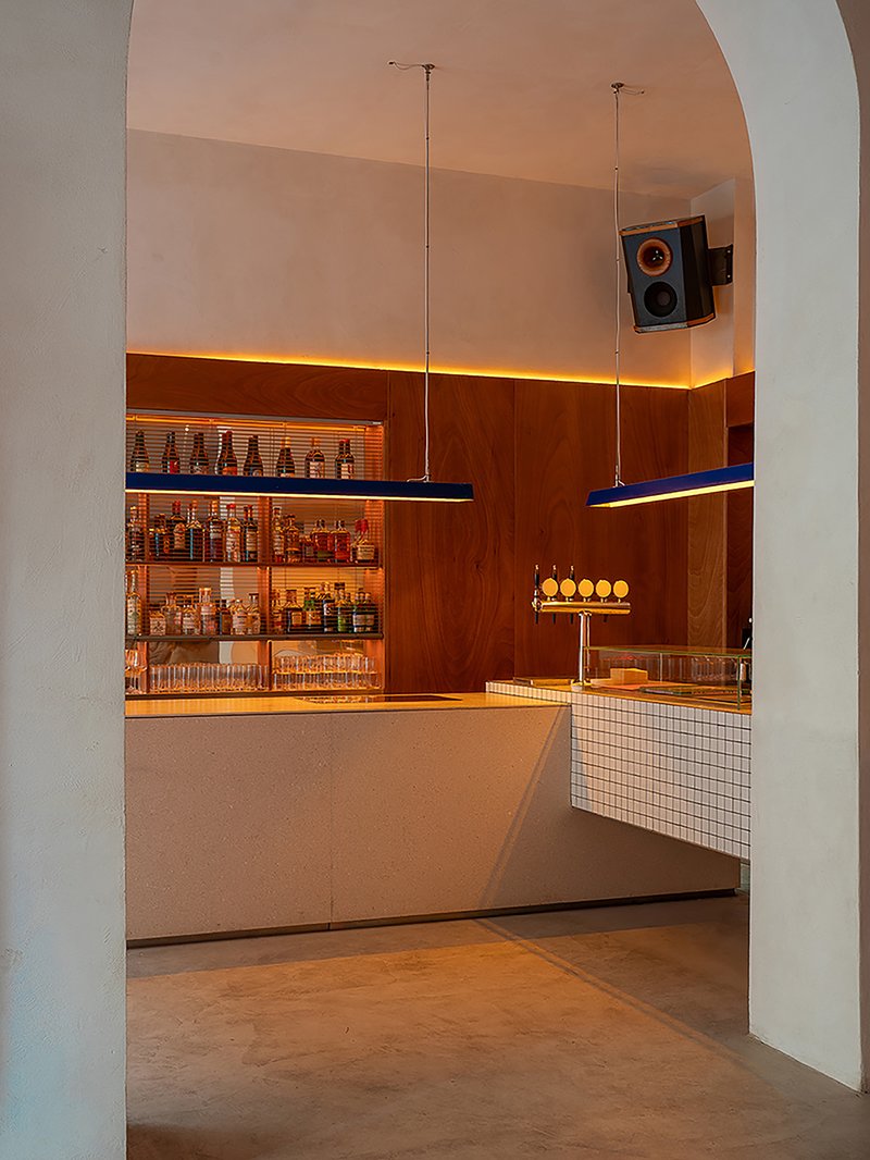 Bar counter with white mosaic tile front, timber paneling, backlit bottle shelves and suspended speakers