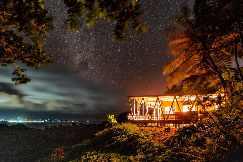 Night view of the illuminated timber pavilion on a hillside beneath the Milky Way
