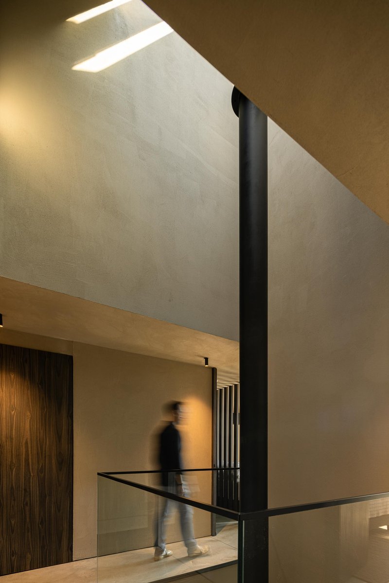Double height hallway with angled skylight illuminating plaster walls and a figure crossing glass balcony