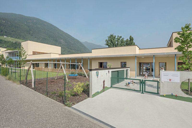 Low-slung building with green entry gates and playground area backed by wooded mountains