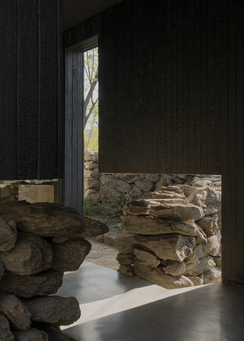 Interior threshold where charred timber wall meets a dry-stacked stone wall bathed in daylight