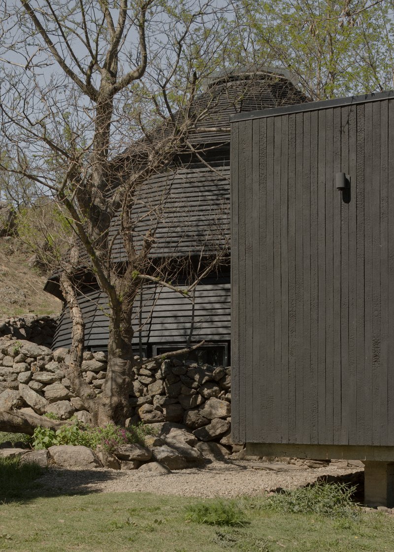 Charred timber facade with curved roof volume and bare tree growing beside dry-stacked stone base