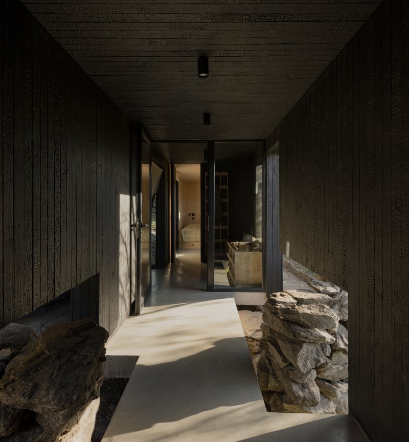 Charred timber corridor with natural light falling across concrete floor and stone walls flanking the passage