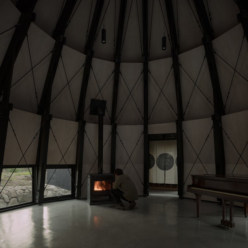 Conical interior with exposed timber ribs as a person crouches beside a glowing wood stove