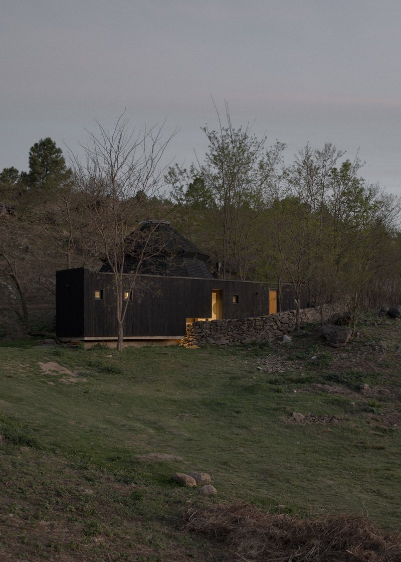 Black timber facade with illuminated window openings at dusk on a hillside terrace