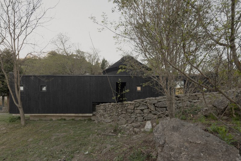 Long black timber box volume beside a dry-stacked stone retaining wall and bare trees