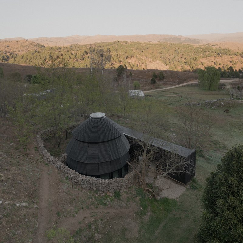 Aerial view of black domed roof and rectangular volume nestled in rolling hills at sunset