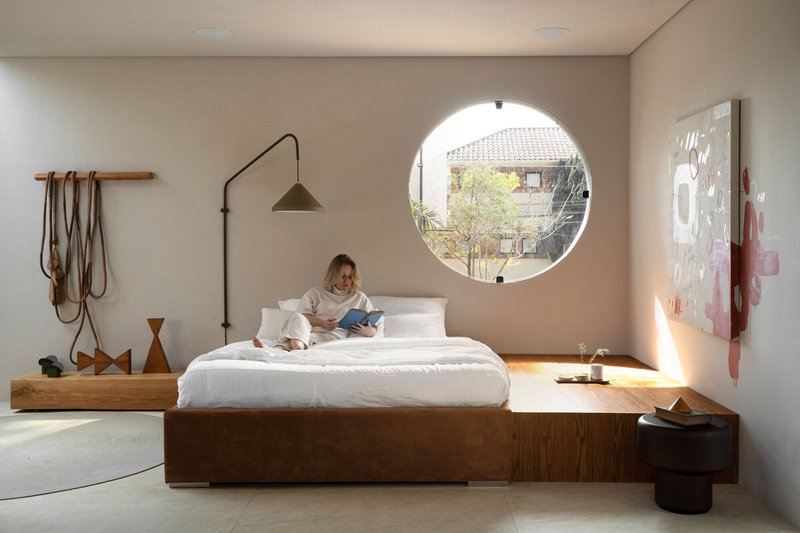 Bedroom with timber platform bed, large circular window and person reading in morning sunlight