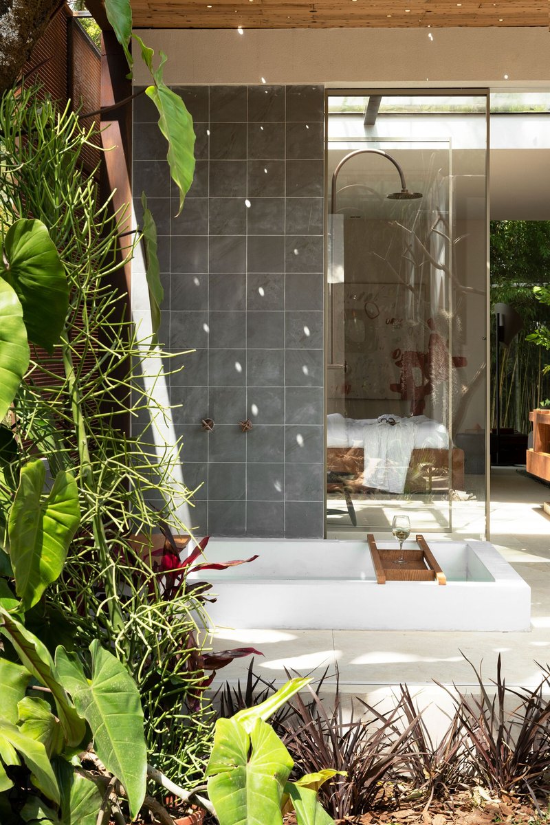 Outdoor shower with white basin beside grey tile wall and perforated metal screen among tropical plants