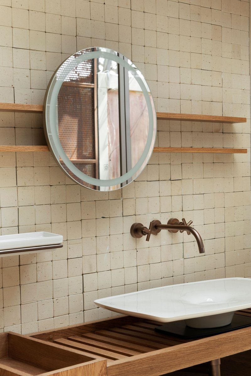 Bathroom vanity with white vessel sink and round mirror against cream tile wall with timber shelves
