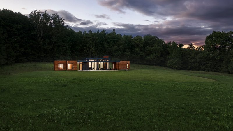 Distant view of the horizontal timber and glass volume set in a meadow at dusk