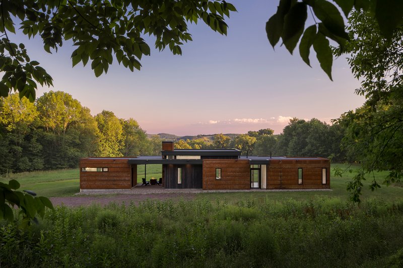 Front facade with horizontal wood cladding and dark glazed sections framed by overhanging tree branches at dusk