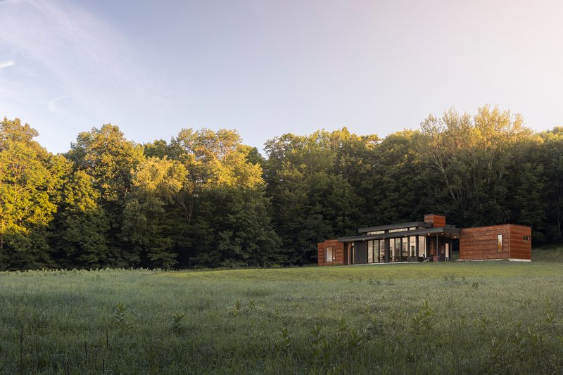 Long view across meadow of the horizontal residence with wood siding and flat roof at sunset