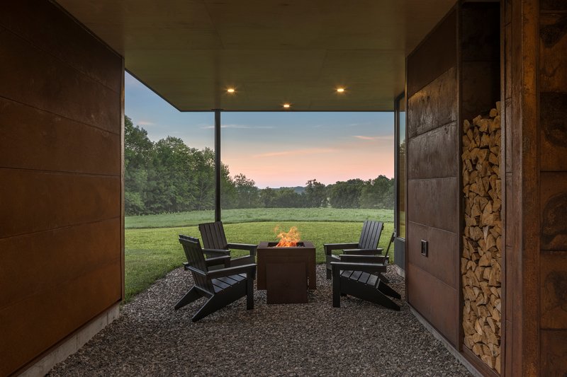 Covered outdoor terrace with fire pit and chairs on gravel looking toward open field at twilight
