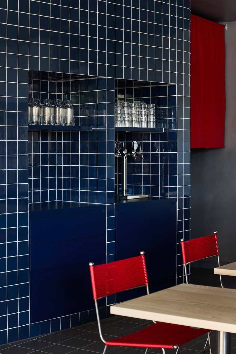 White-grouted blue tile wall with recessed shelving, chrome tap, and red canvas chairs at a table