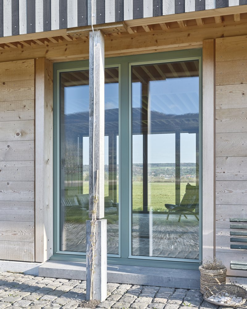 Green-framed glass doors in a timber and concrete facade reflecting the landscape under an exposed rafter ceiling