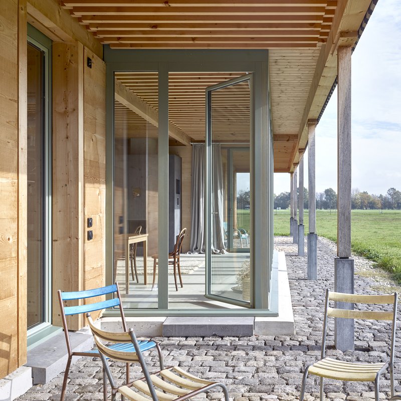 Covered porch with exposed timber rafters and folding metal chairs beside floor-to-ceiling glazing on gravel