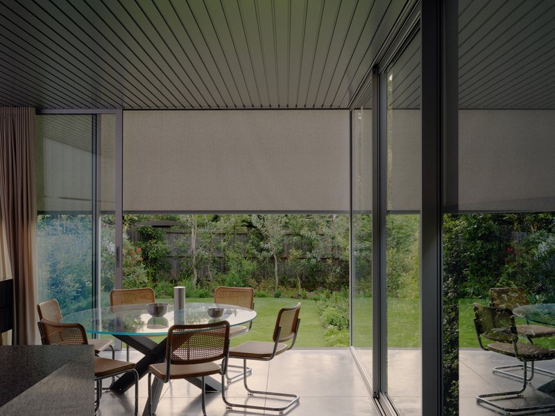 Interior dining space with floor-to-ceiling glazing overlooking garden and retractable roller shades