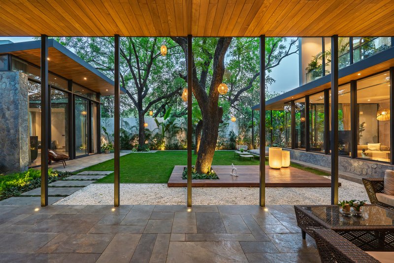 View through timber-framed glazing into a central courtyard with a mature tree and surrounding pavilions at dusk