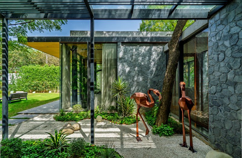 Steel pergola framing a view of the tree trunk wrapped in stone wall with gravel and planted ground cover