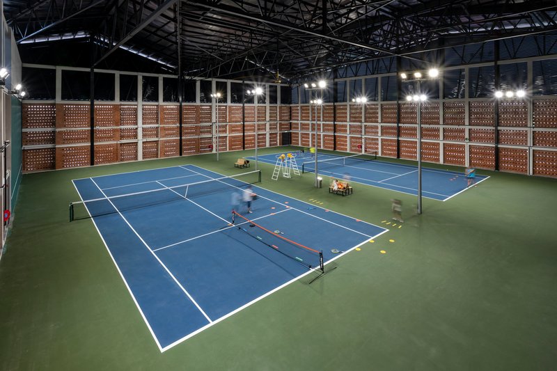 Interior view of two blue tennis courts surrounded by perforated brick screens under an exposed steel truss roof