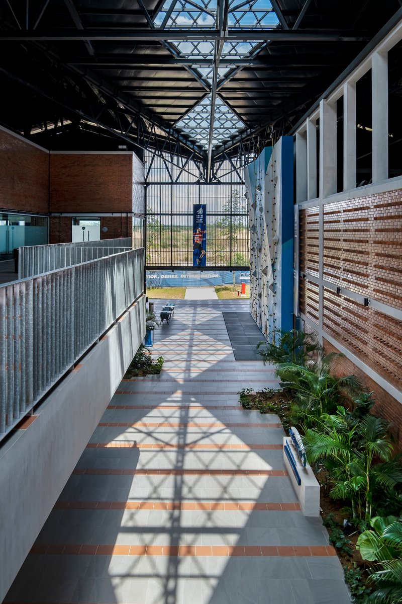 Interior corridor with exposed steel trusses, patterned shadows, planted beds, and glazed end wall