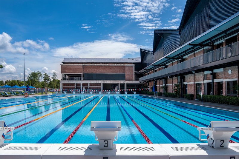 Olympic-sized swimming pool with lane markers, starting blocks, and brick pavilion under scattered clouds