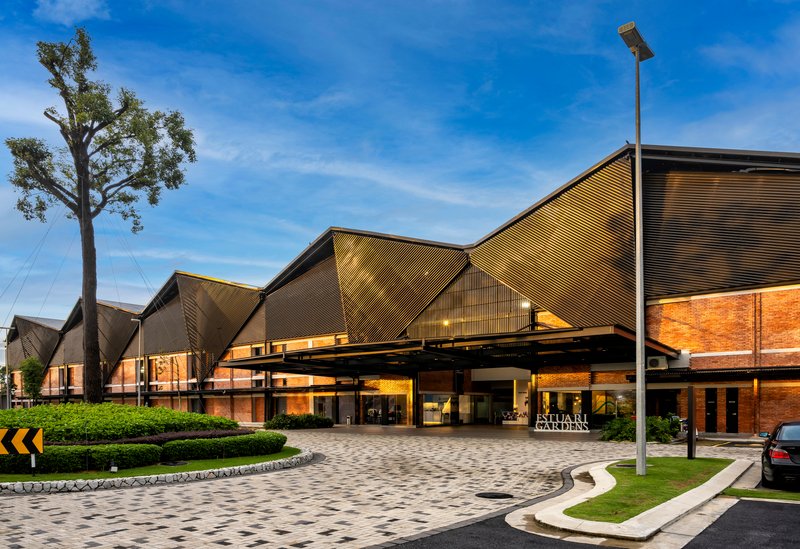 Entry canopy with folded metal roof planes over a paved forecourt with mature trees