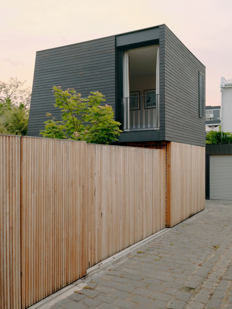 Street view of dark horizontal cladding above a vertical timber fence with trees behind