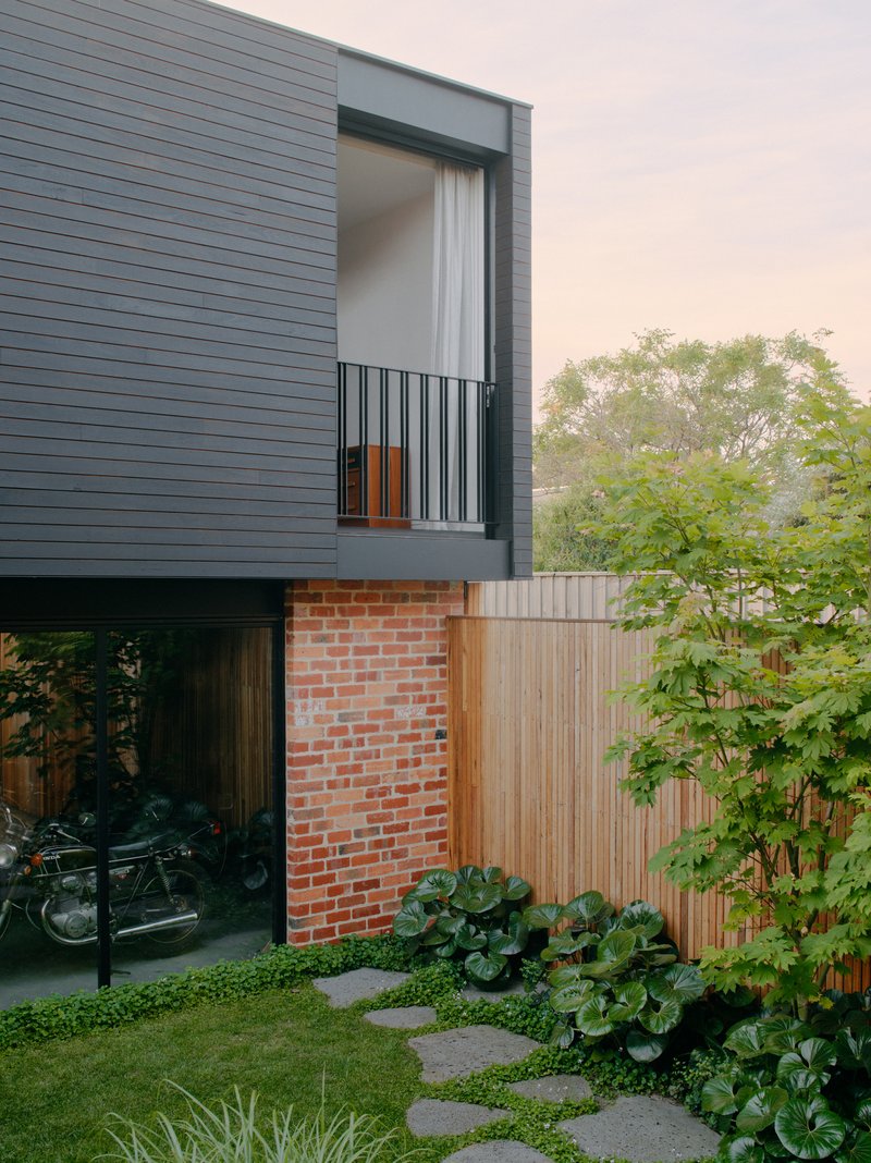 Rear elevation with cantilevered dark cladding over brick base, timber fence and garden bed at dusk