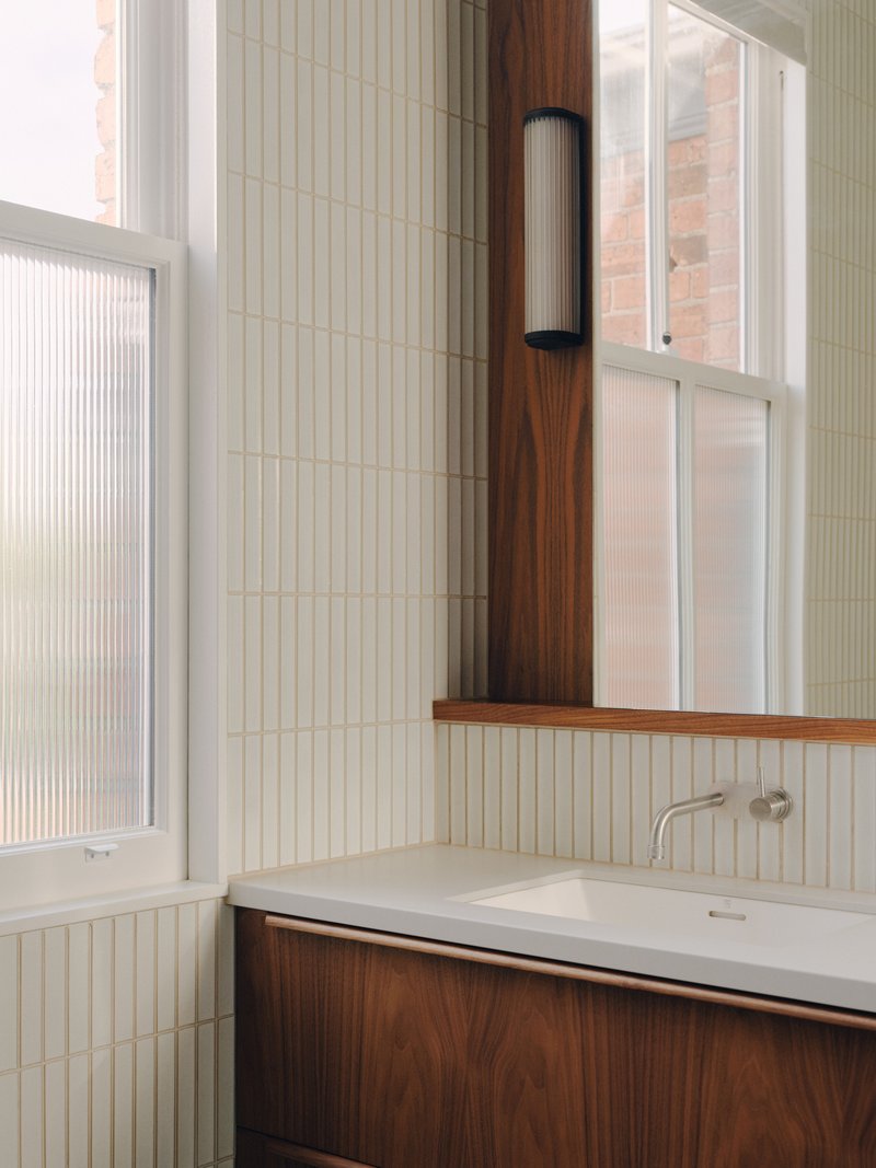 Bathroom vanity corner showing vertical tile, walnut shelf, and cylindrical wall sconce near window