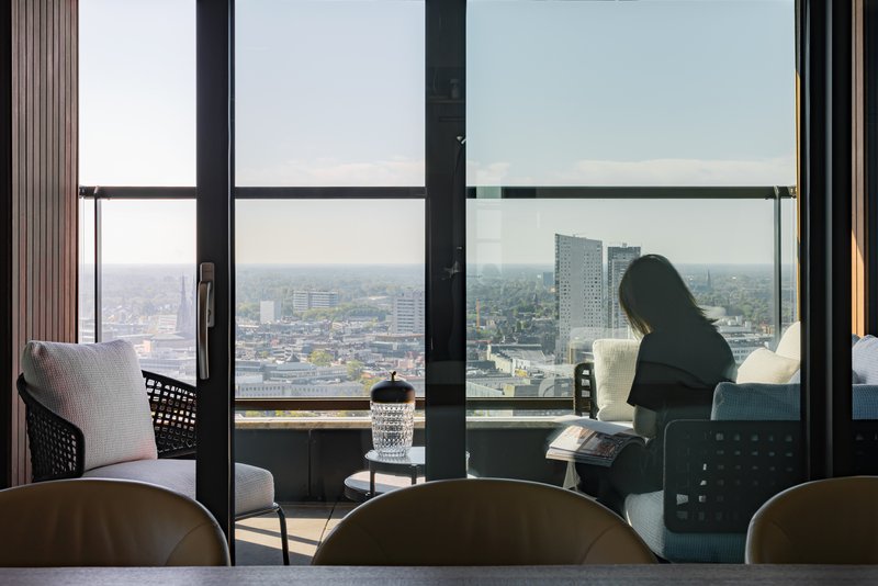 Interior view through floor-to-ceiling glazing with a figure reading on the balcony overlooking the cityscape